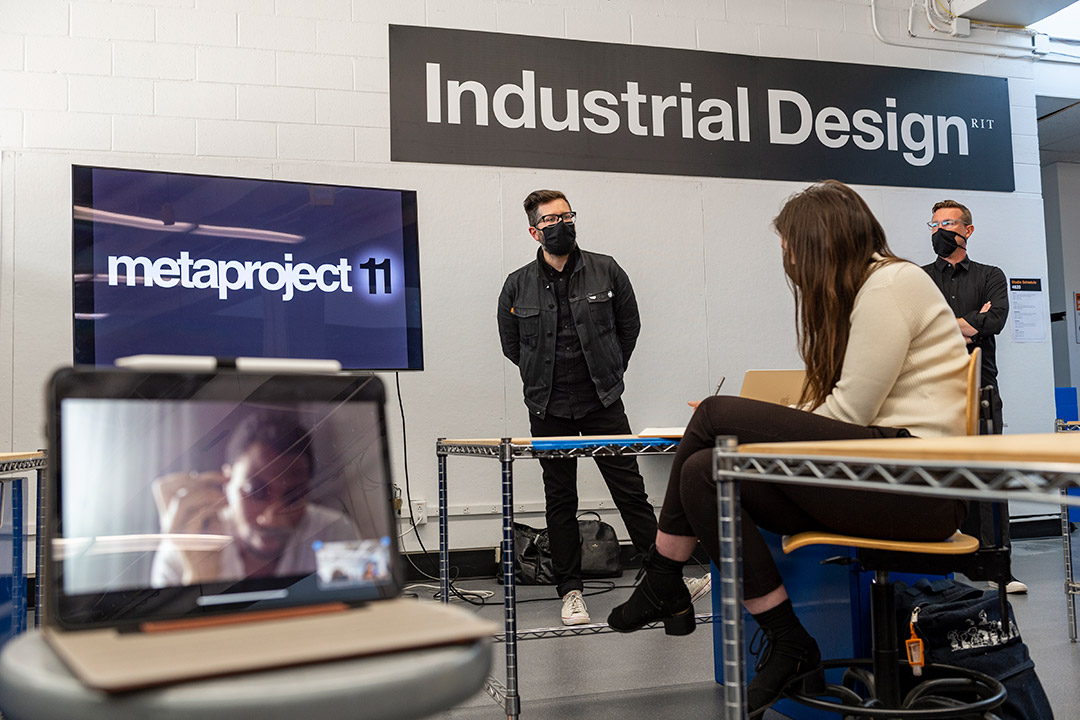 presenters standing at front of class with students in person and viewing from a laptop.