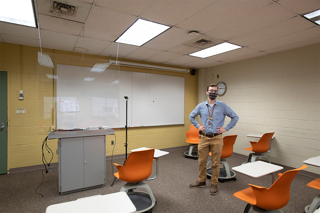 student worker standing in classroom with plexiglas hanging from ceiling.