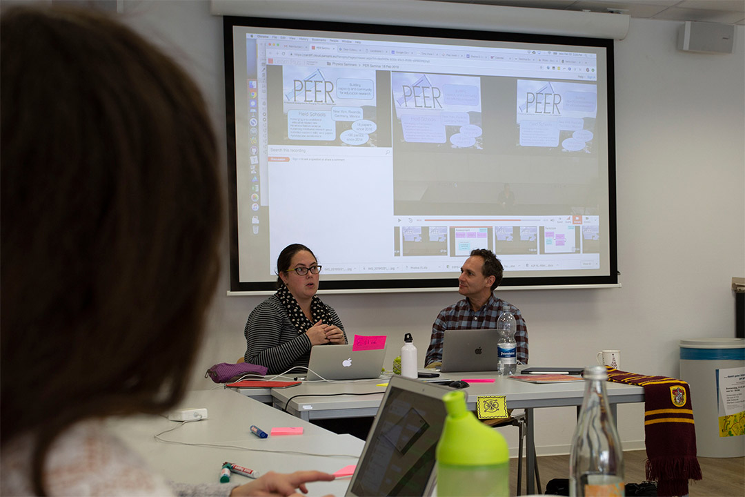 two presenters sitting at a table with laptops and projector screen behind them.