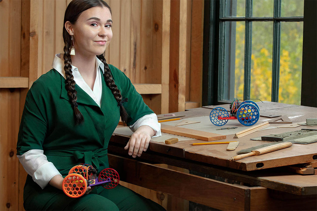 woman sitting at wooden desk holding a small, colorful robotic vehicle.