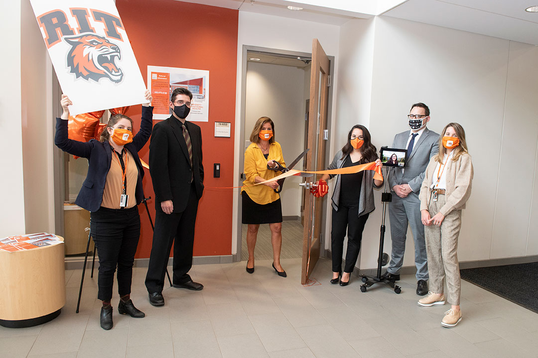 six people celebrate opening of a clinic by cutting a ribbon with oversized scissors.