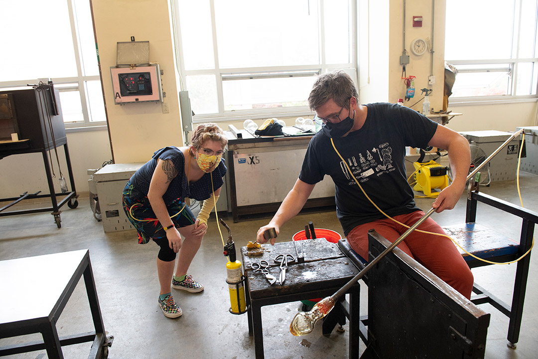 student observing as professor blows glass with hose and tube.