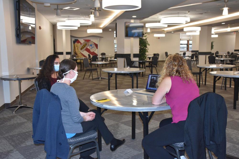 three employees wearing face masks sitting at a round table watching a presentation on a laptop.