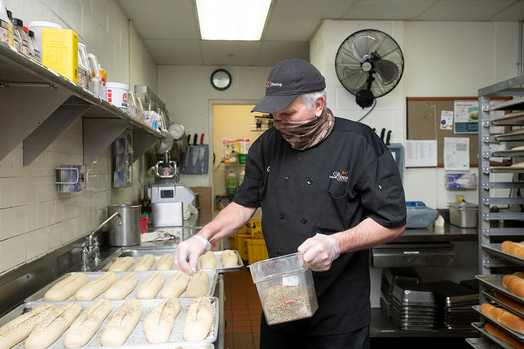 food service worker sprinkling sesame seeds onto loaves of bread dough.