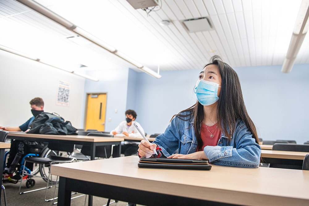 students wearing face masks and sitting one to a table in a classroom.