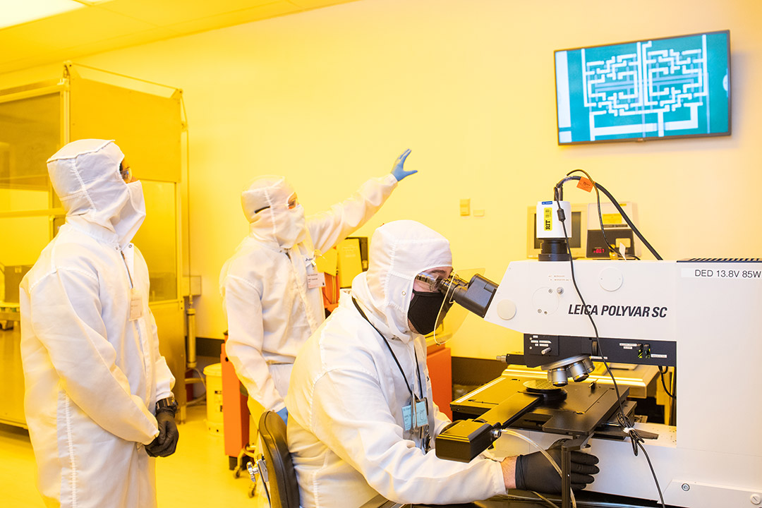 researchers wearing clean suits analyzing a magnified view of an integrated circuit.