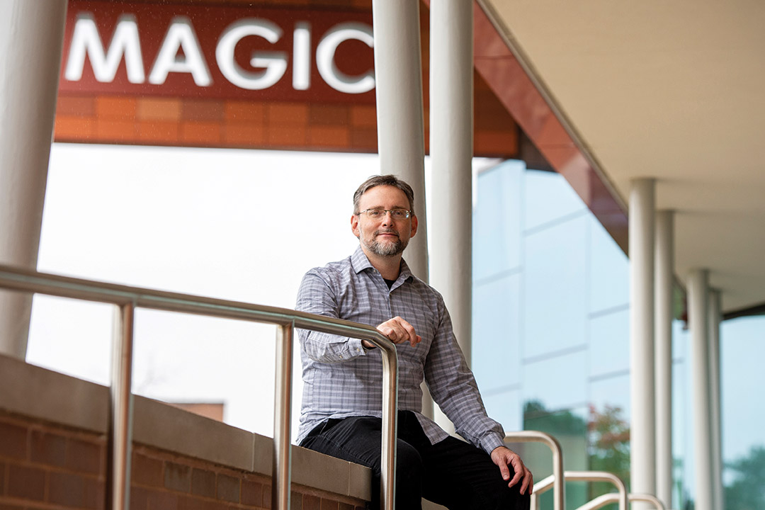 professor posing outside with building sign that says MAGIC in the background.