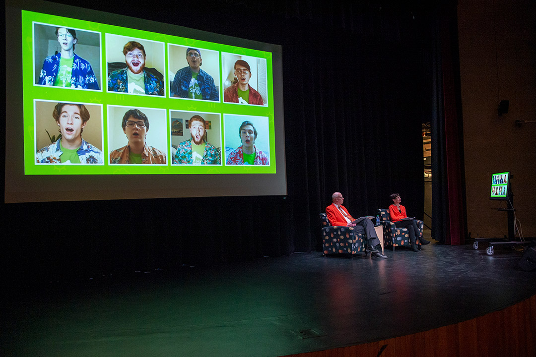 two people sitting on stage watching a TV screen with the image also projected on large screen behind them.