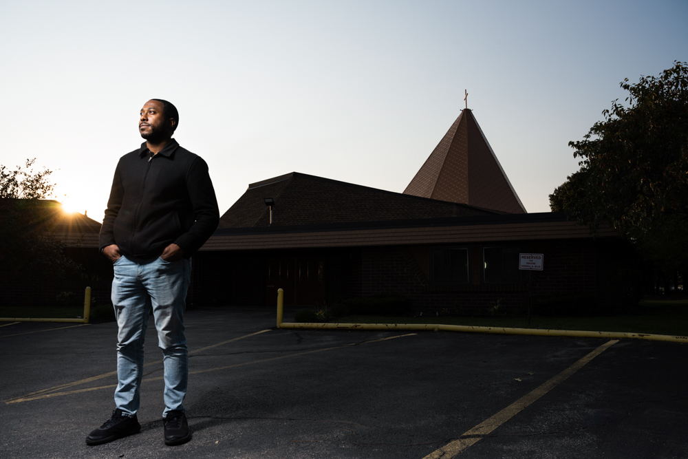 A man poses for a portrait in the parking lot, with a church in the backdrop.