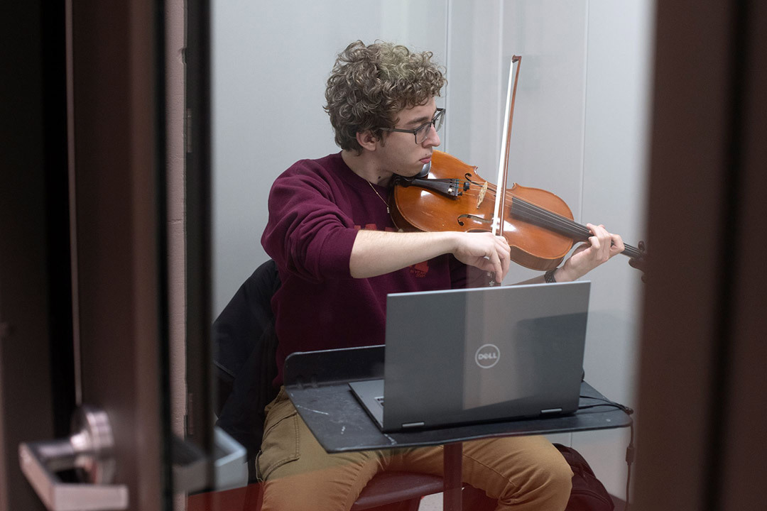 A student playing a viola, while sitting in front of a laptop.