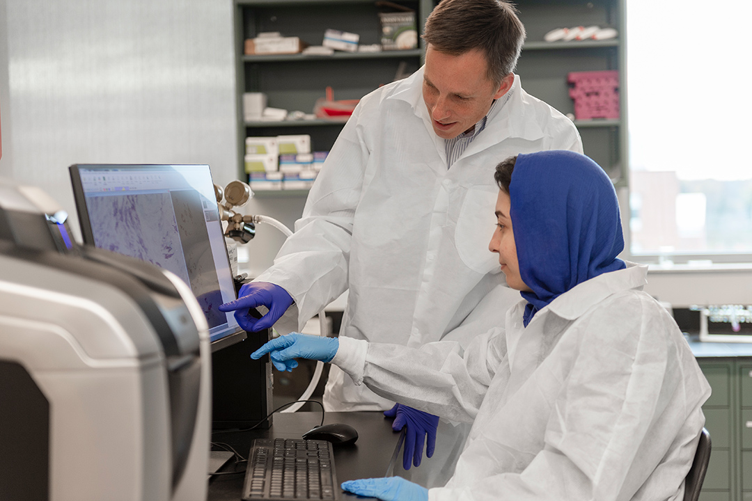 professor and student wearing lab coats and gloves pointing to computer monitor.