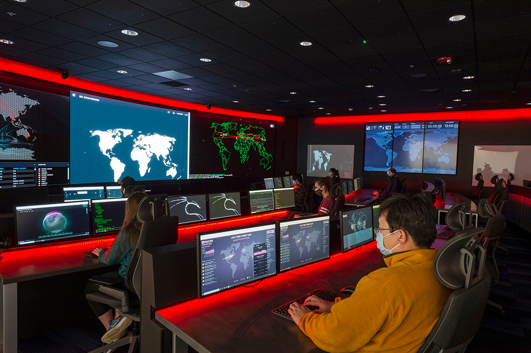 people on computers in rows in a large, dark room with TV and projector screens.