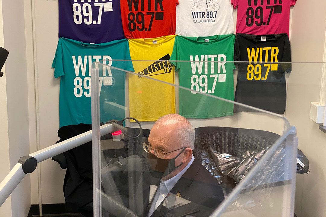 Man wearing a facemask sits in radio studio talking into microphone.