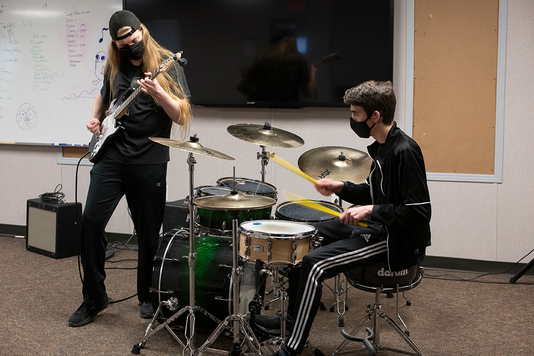 student playing the electric guitar next to student playing drums.