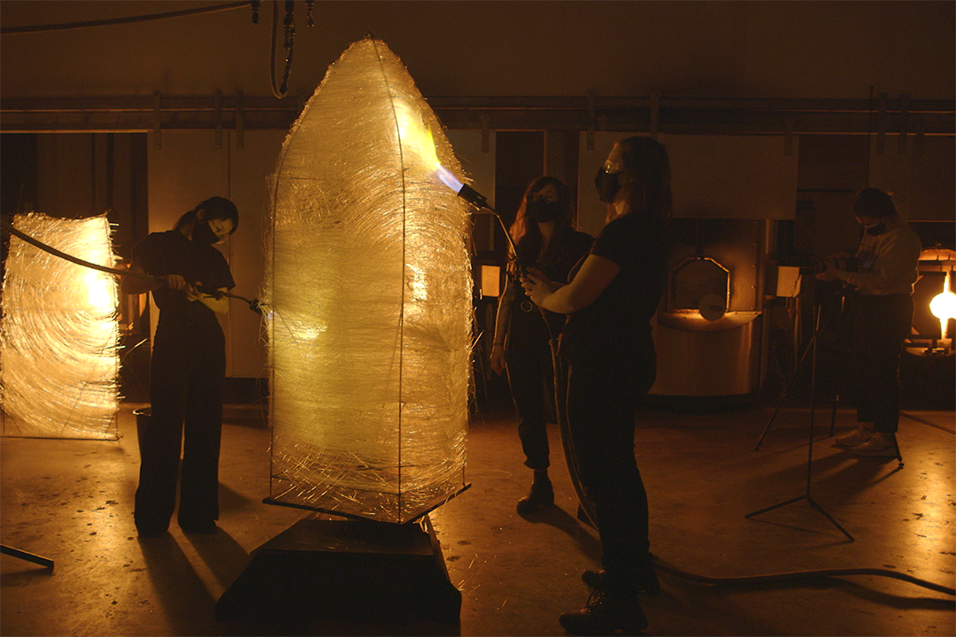 A group of artist take flame torches to a large, woven sculpture.