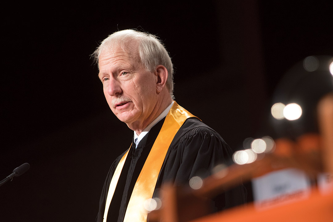 alumnus wearing graduation regalia speaking at podium.