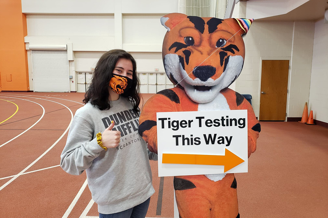 student poses with cardboard cut-out of tiger mascot with sign Tiger Testing This Way.