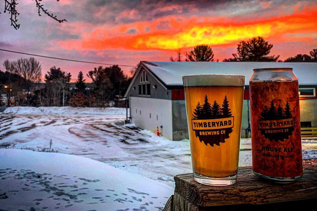 can and glass of beer in foreground of snowy scene with brewery in background.
