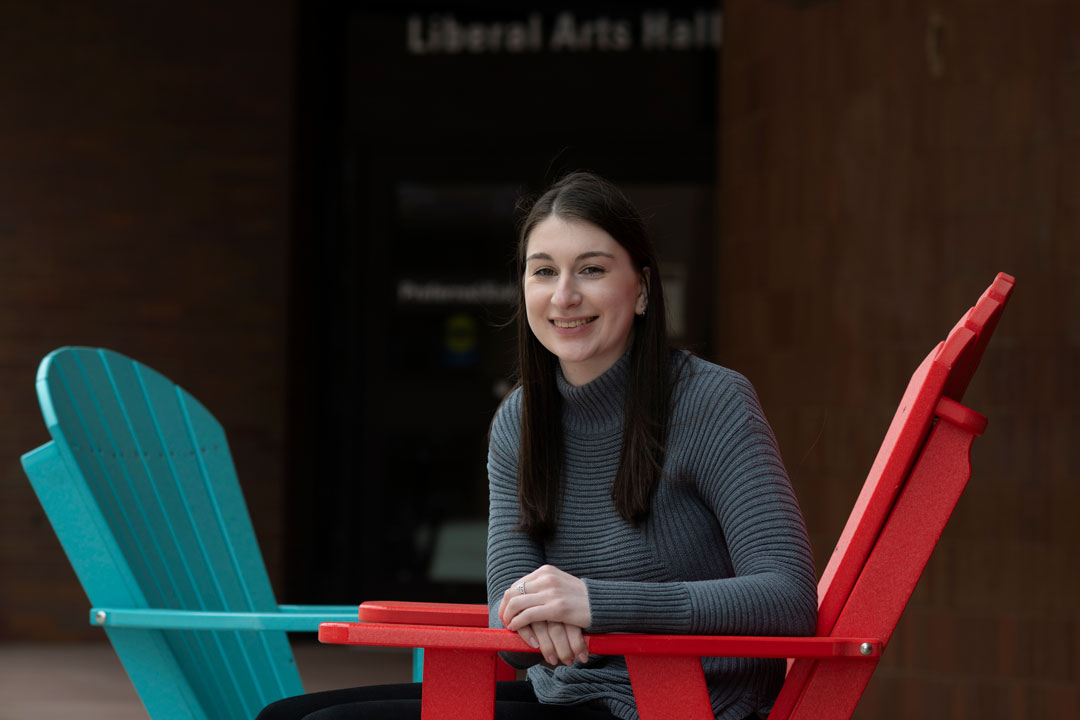 A student sits in a red chair in front of a brick building.