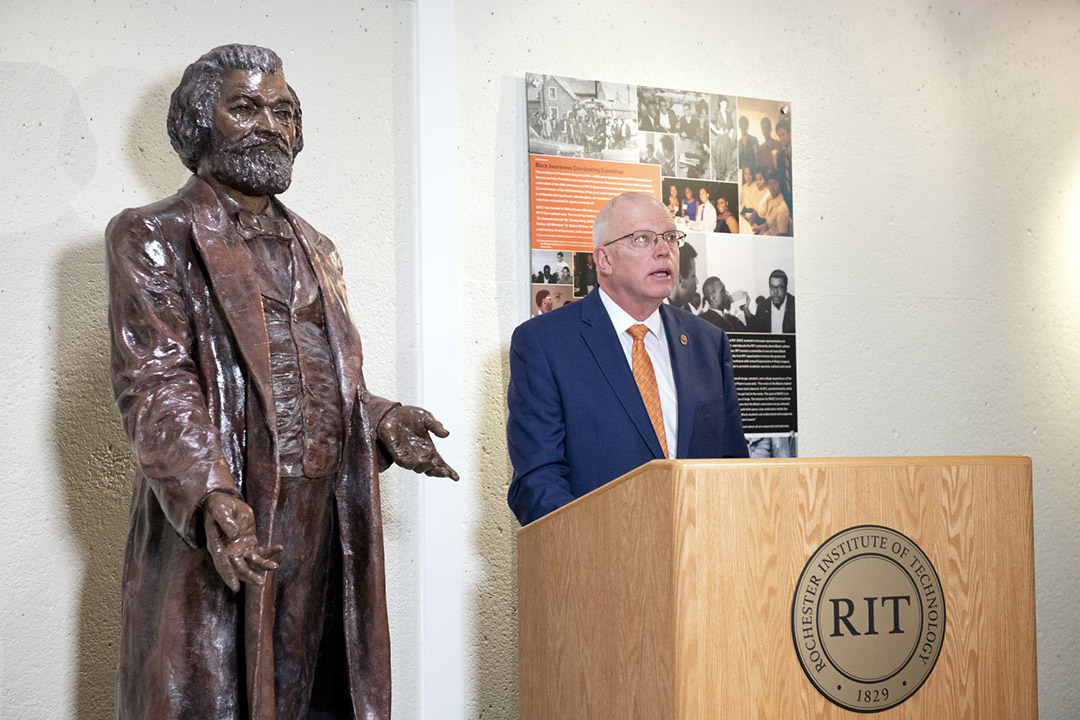 RIT President David Munson standing at a podium next to a statue of Frederick Douglass.