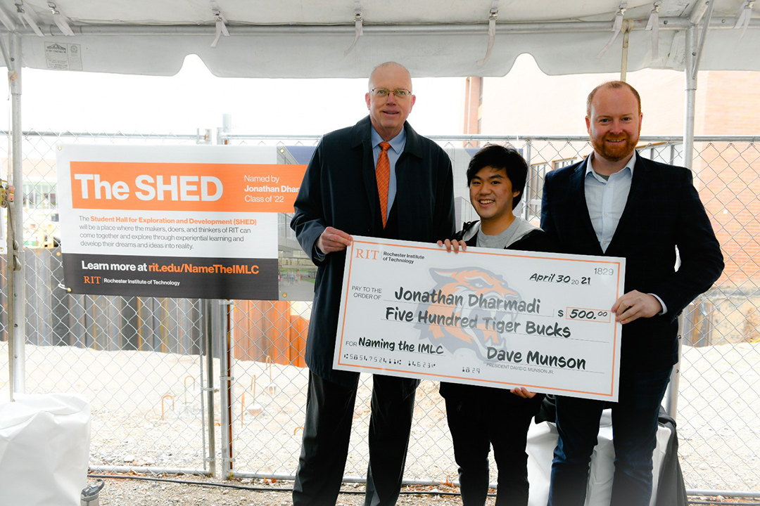 Three men standing with a giant check for 500 dollars. The sign behind them says The SHED.