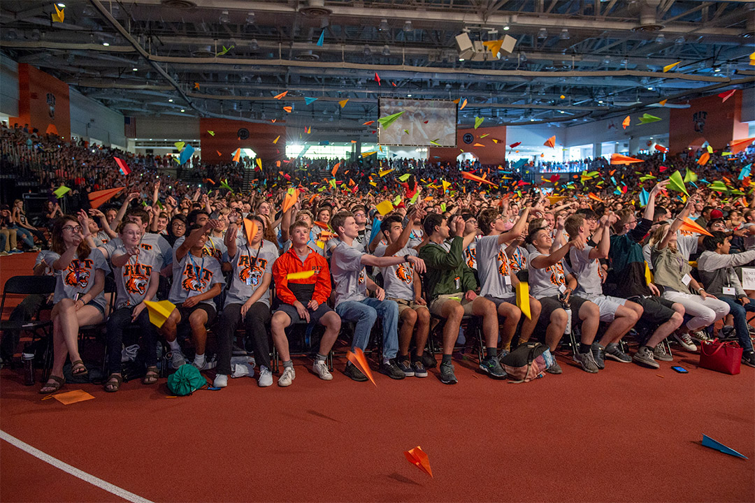 crowd of seated students throwing colorful paper airplanes.