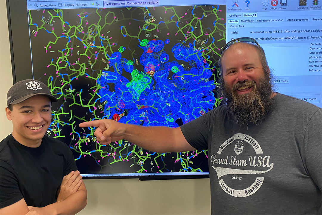 student and professor standing in front of a projection of a computer screen showing colorful depictions of proteins and bacteria.