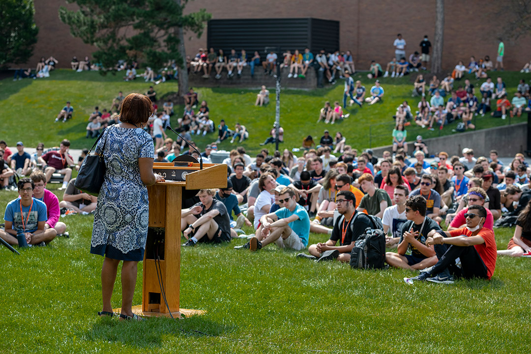 groups of students sitting on the grass listening to a speaker at a podium.