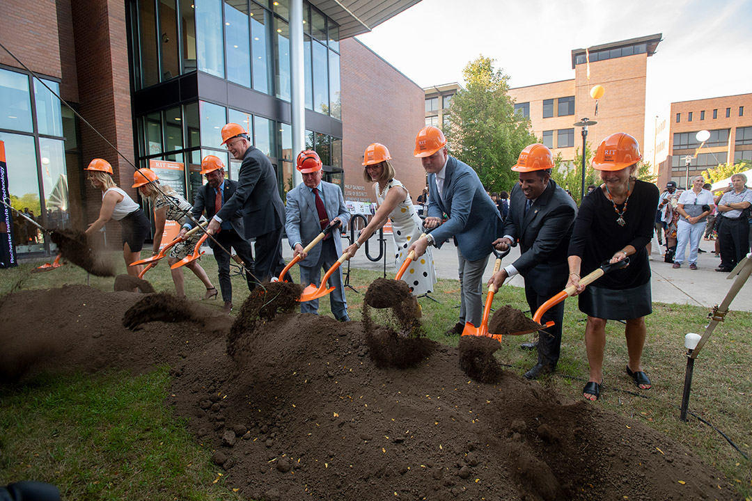 nine people wearing hard hats using shovels to dig a small portion of dirt in a ceremonial groundbreaking.