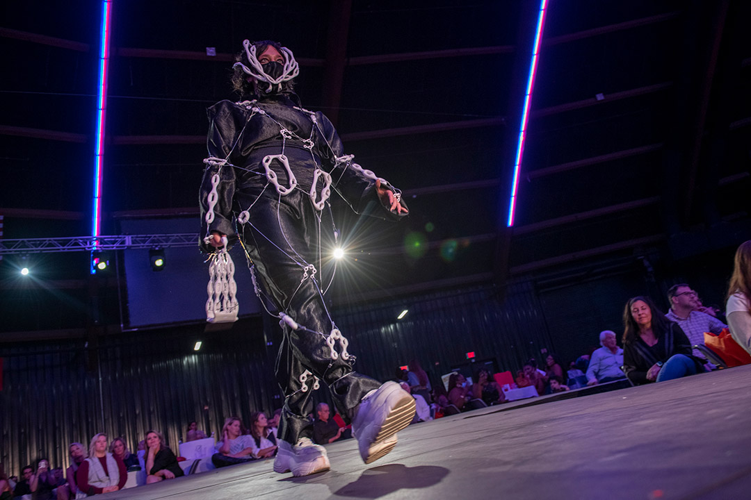 student walking a runway wearing a piece that features black fabric and white chain-like structures.
