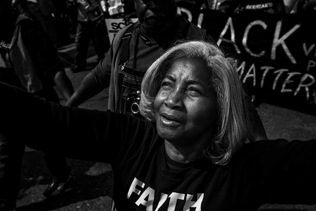 black and white photo of a woman with her arms outstretched and a Black Lives Matter sign in the background.