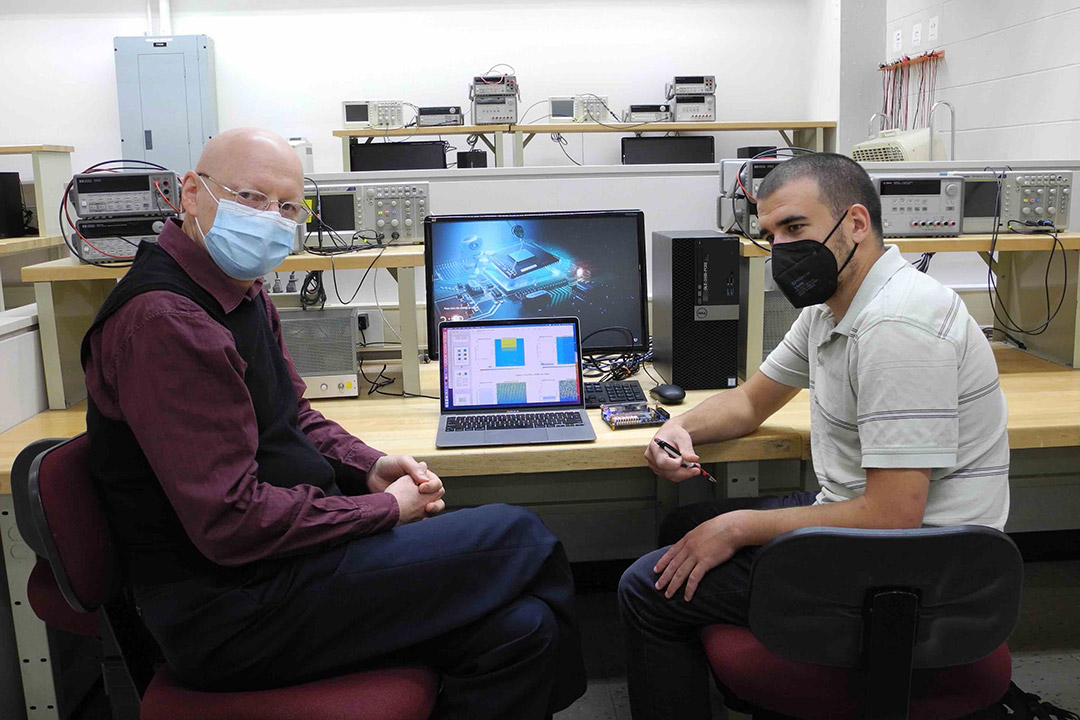 two researchers wearing masks and sitting next to a computer setup.