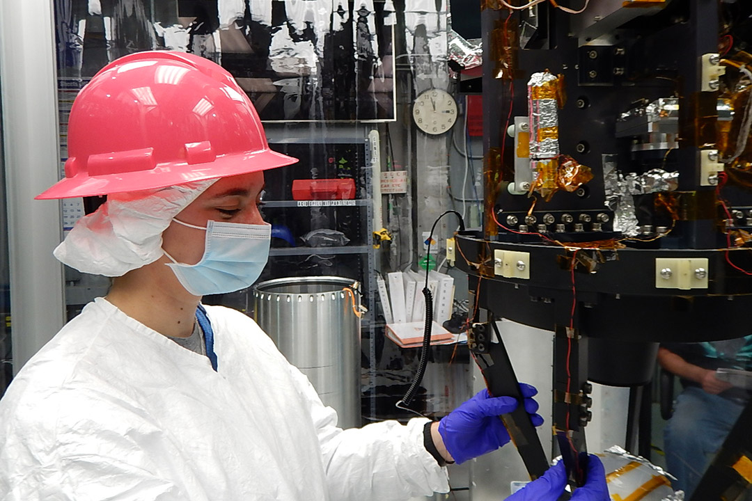 student wearing hard hat and clean suit working on rocket equipment.