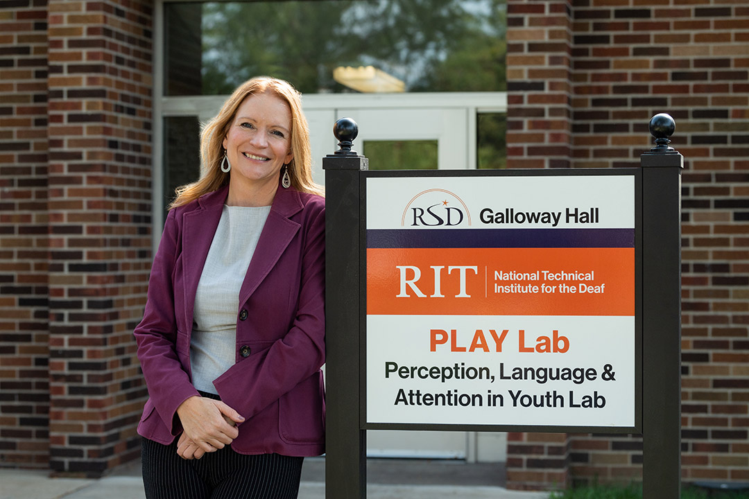 researcher standing outsite next to a sign that reads Play Lab, perception, language and attention in youth lab.