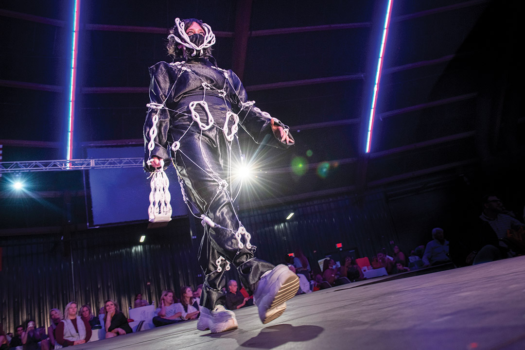 student walking a runway wearing a piece that features black fabric and white chain-like structures.