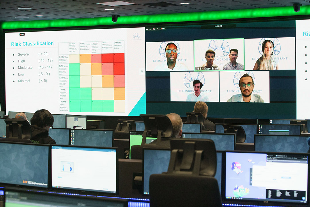 students sitting at rows of computers with several large screens projecting charts and team members' faces.