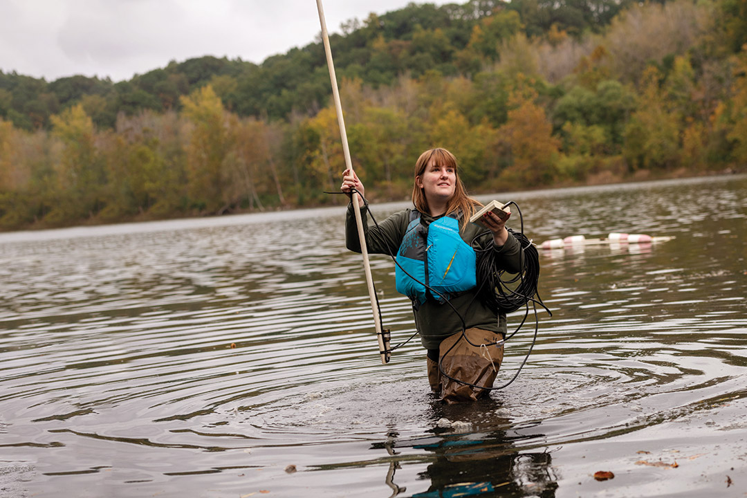 student research in waders in a lake with a pole and a measuring device.