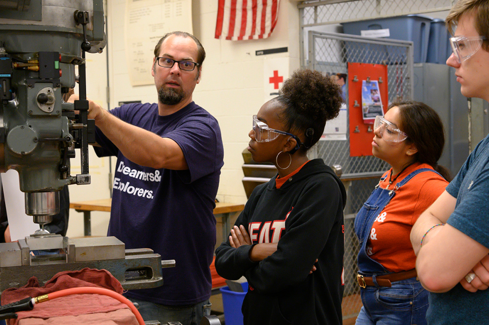 staff member shows three students a drill press.