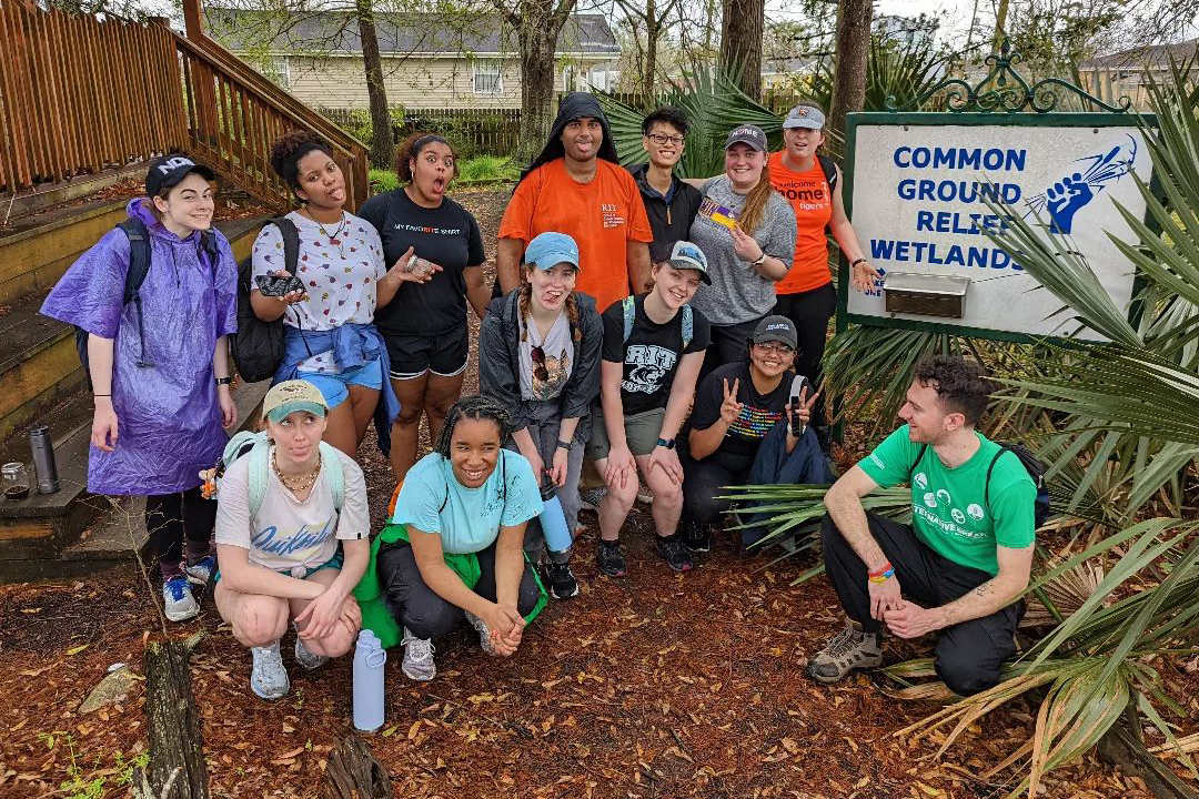 group of 13 students poses outdoors next to sign for Common Ground Relief Wetlands.