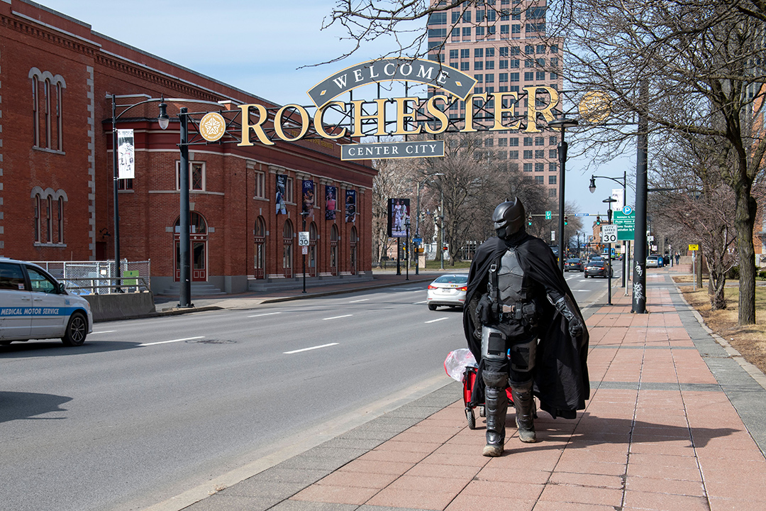a student dressed as Batman walks past the Welcome to Rochester sign.