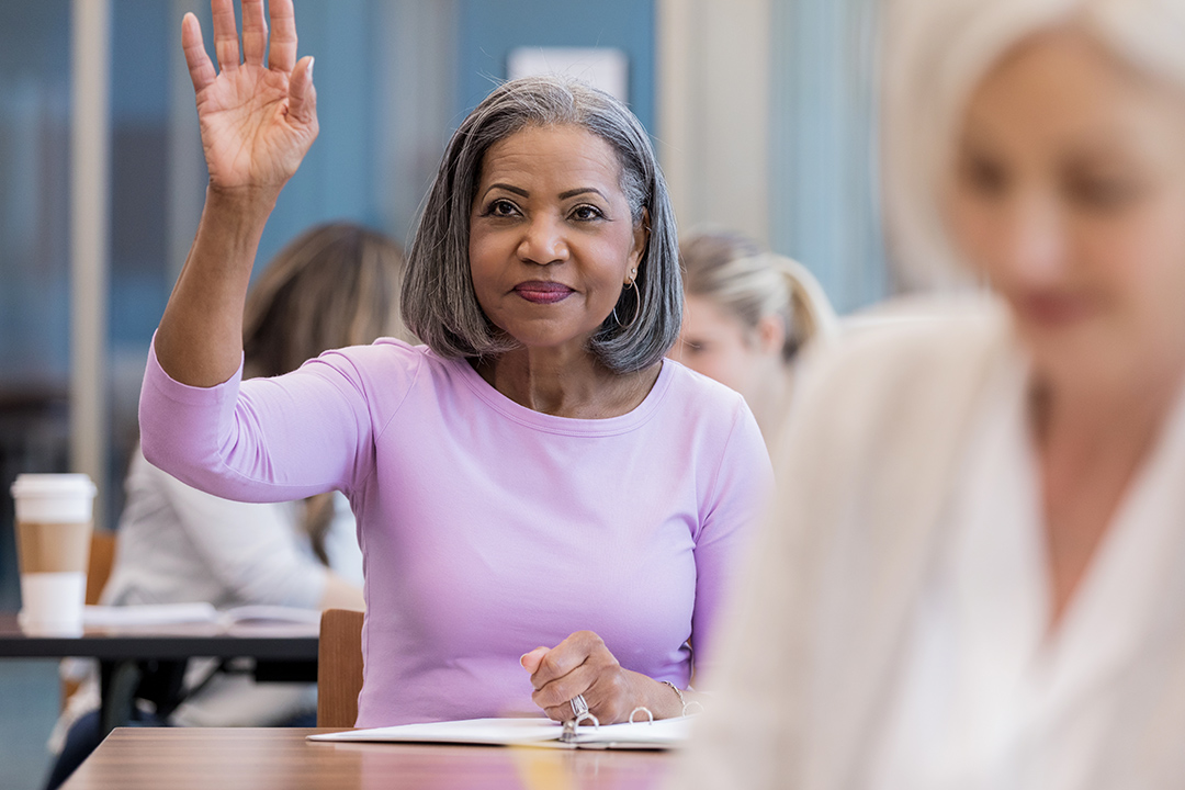 woman with gray hair in a classroom setting raising her hand.