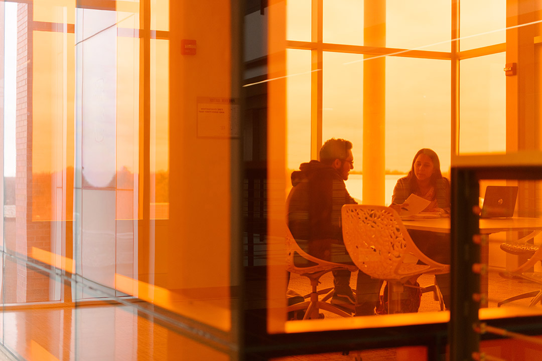 two students working in a room surrounded by orange windows.