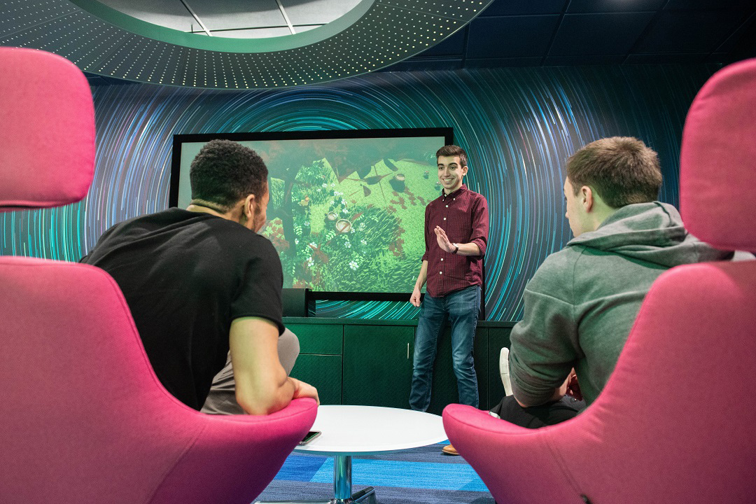 student presenting video game to two people seated in red chairs.