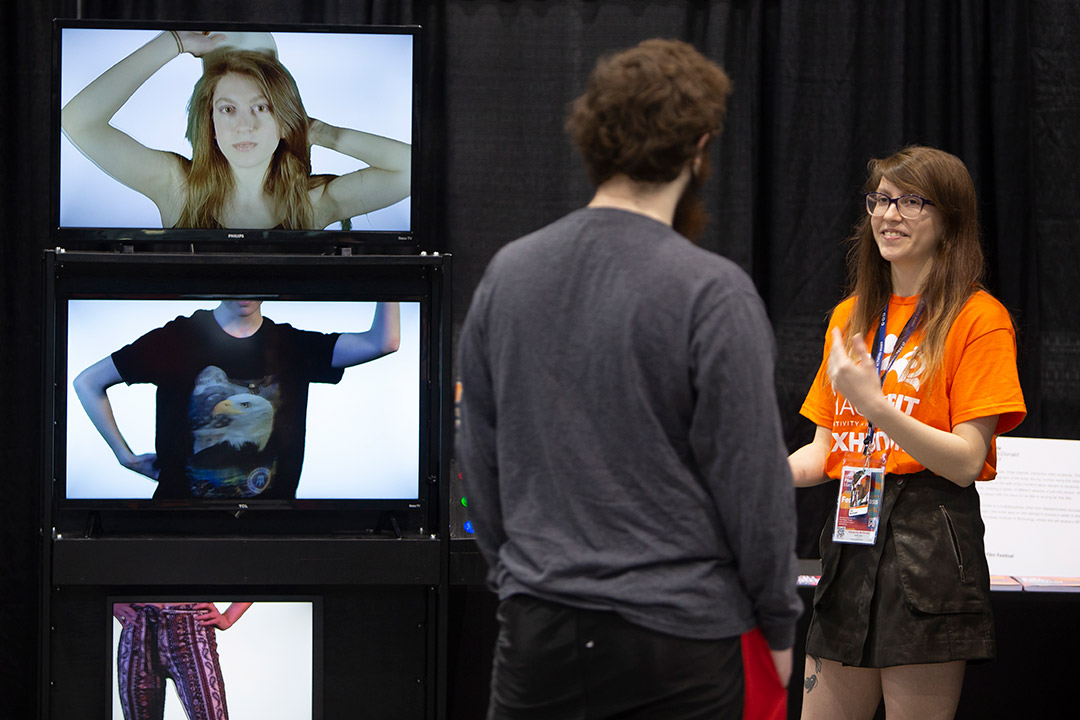 two students talking next to three stacked TV screens showing a person's head, torso, and legs.
