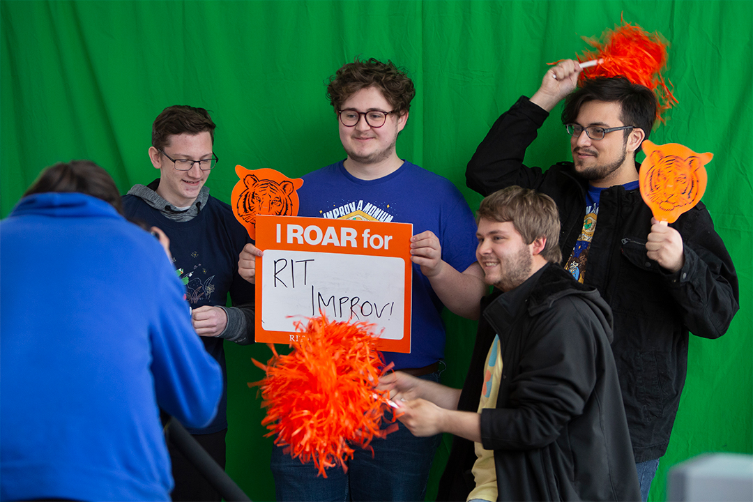 Four male students pose for a camera in front of a green screen and the one in the center holds a sign that said, I ROAR for RIT Improv!
