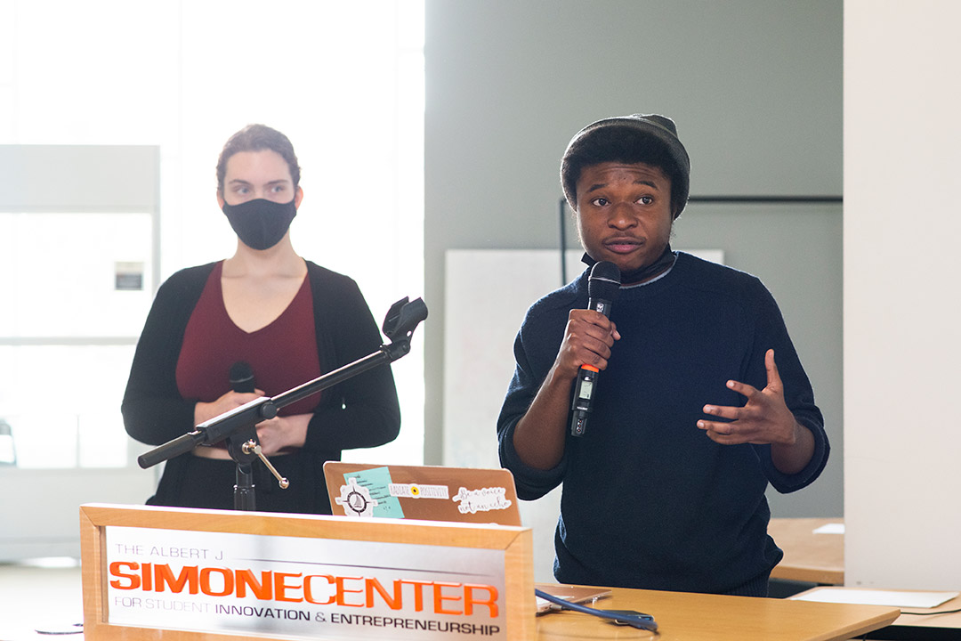 student speaking into a microphone at a podium with another student standing in the background.