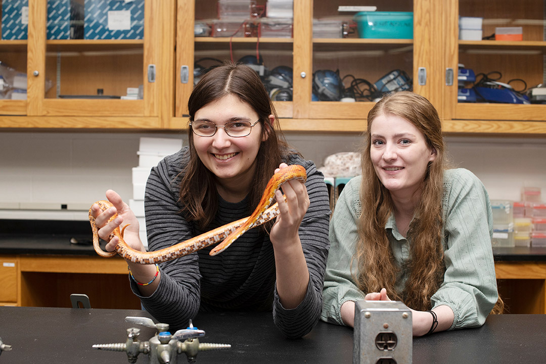 two students in a science lab, and one is holding a small orange snake.
