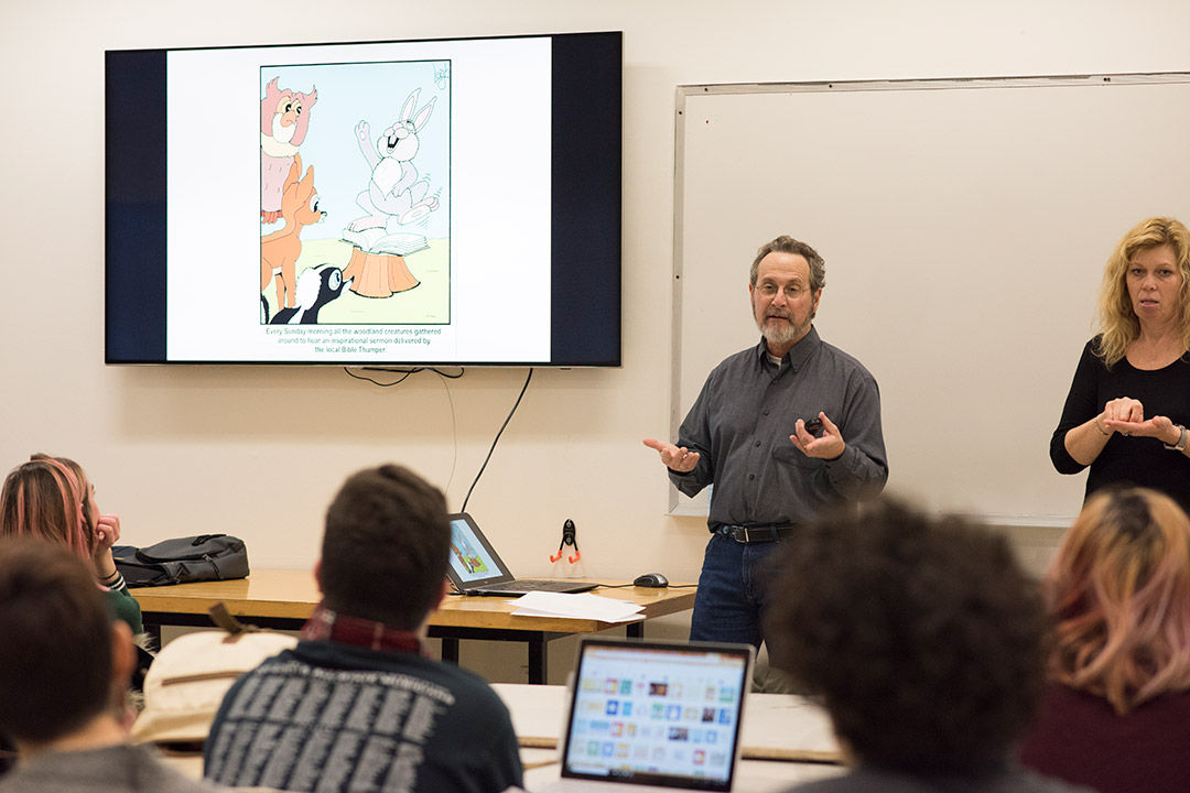 man standing in front of a classroom giving a presentation, with a screen showing a cartoon panel.