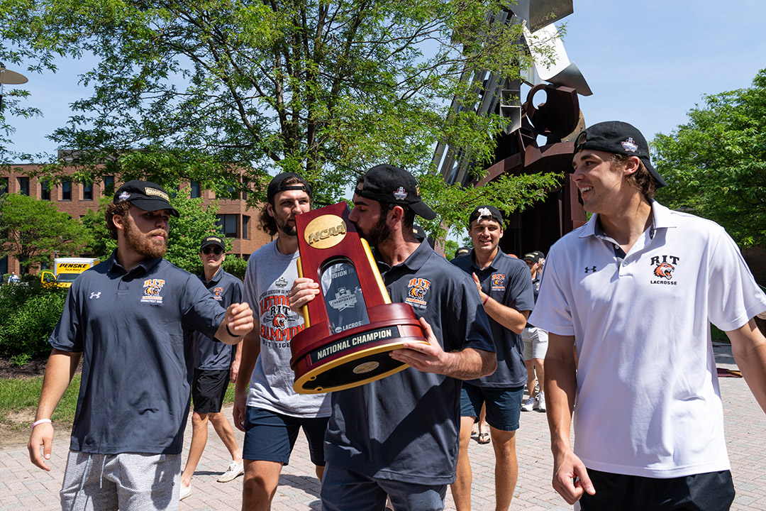 men's lacrosse team members walking while one kisses the championship trophy.