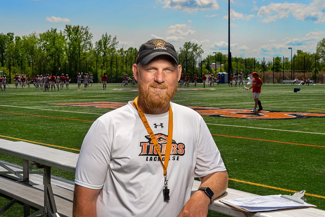 'lacrosse coach standing next to the bleachers as the men's lacrosse team practices in the background.'